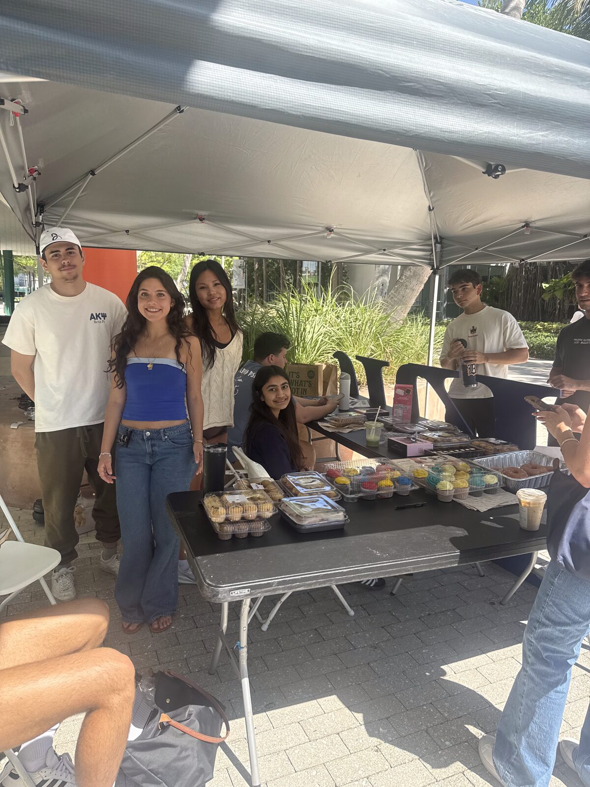 Brothers at a bake sale table under a tent on campus